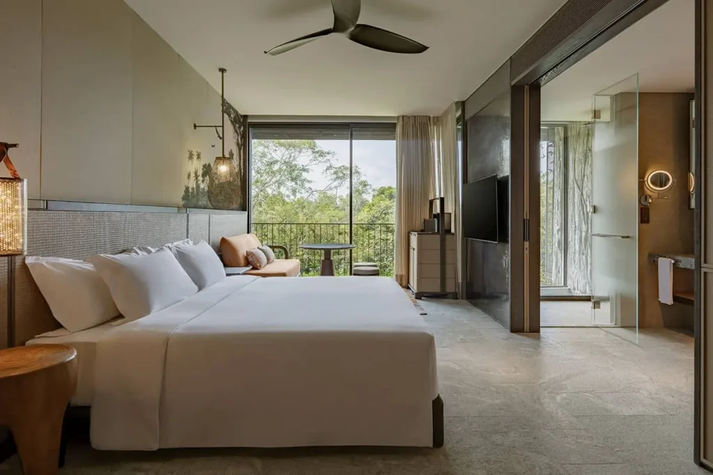 This contemporary hotel suite features a minimalist design with neutral tones, floor-to-ceiling windows, and a private balcony overlooking a lush tropical forest. The interior includes a large bed with crisp white linens, sleek stone flooring, and integrated lighting that enhances the warm, organic atmosphere.