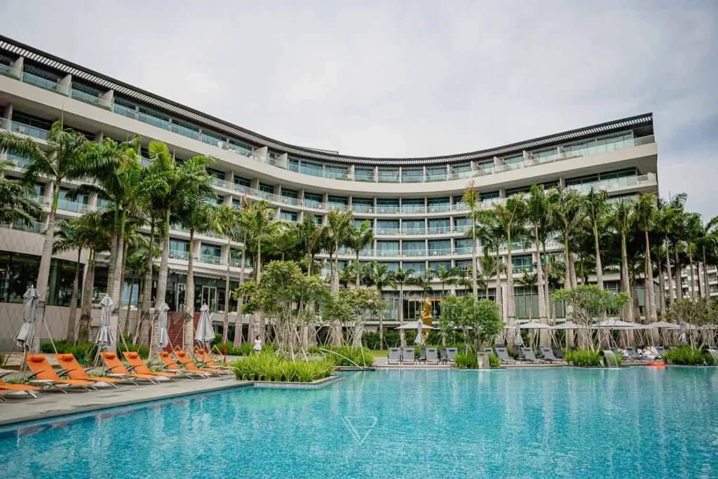 This image captures the expansive outdoor pool area of the W Singapore - Sentosa Cove, featuring a curved modern hotel facade lined with private balconies. The tropical setting is highlighted by numerous palm trees, vibrant orange lounge chairs, and clear blue water under a bright sky.