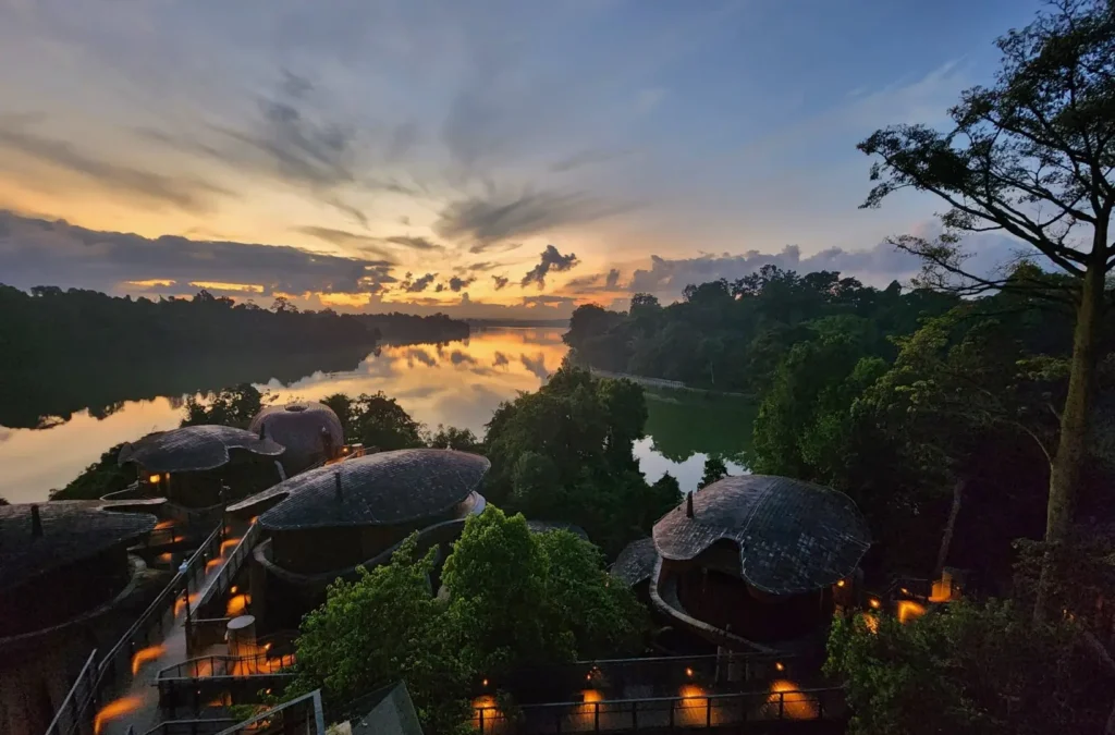 A breathtaking sunset casts a warm golden glow over a serene river, which perfectly reflects the dramatic clouds and surrounding lush greenery. In the foreground, unique organic-shaped villas with illuminated walkways are nestled into the forest hillside, overlooking the tranquil water.