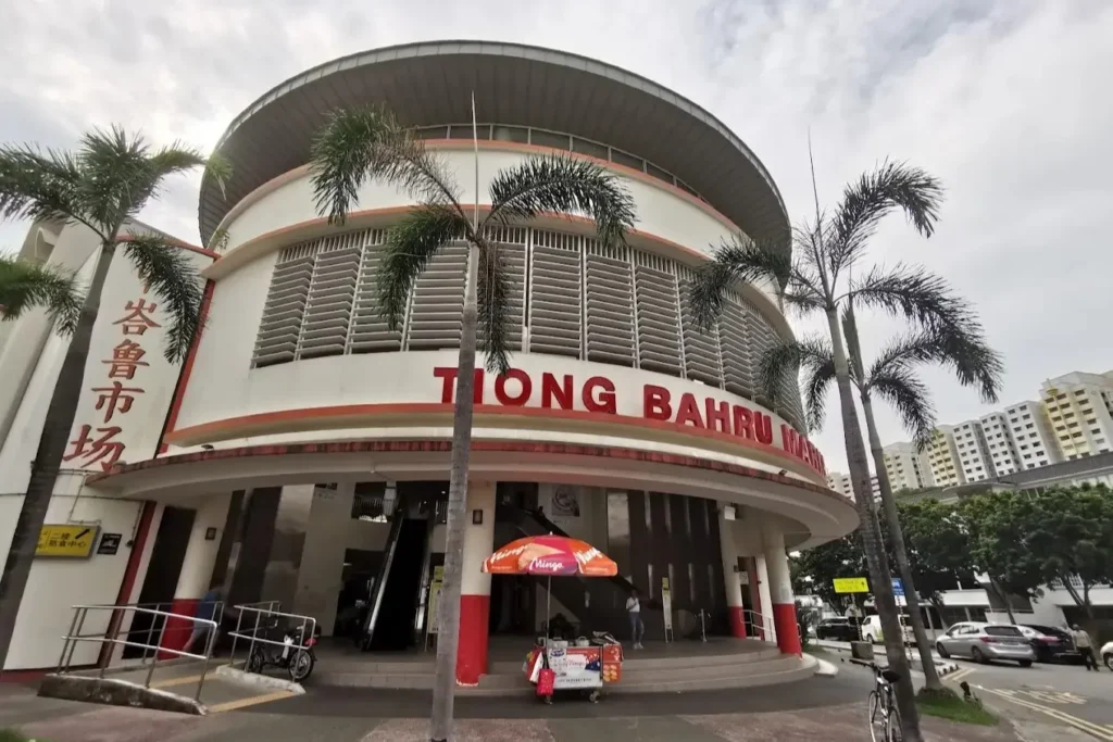 The image shows the curved, modern exterior of the Tiong Bahru Market in Singapore, featuring large red lettering and a distinctive rounded roof. Tall palm trees and a small ice cream cart stand in front of the building's entrance.