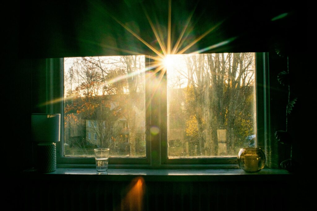A golden sunburst streams through a window, casting a warm glow over a sill adorned with a lamp, a glass of water, and a candle. The bright light illuminates a view of autumn trees and a distant yellow building, creating a cozy, serene atmosphere.