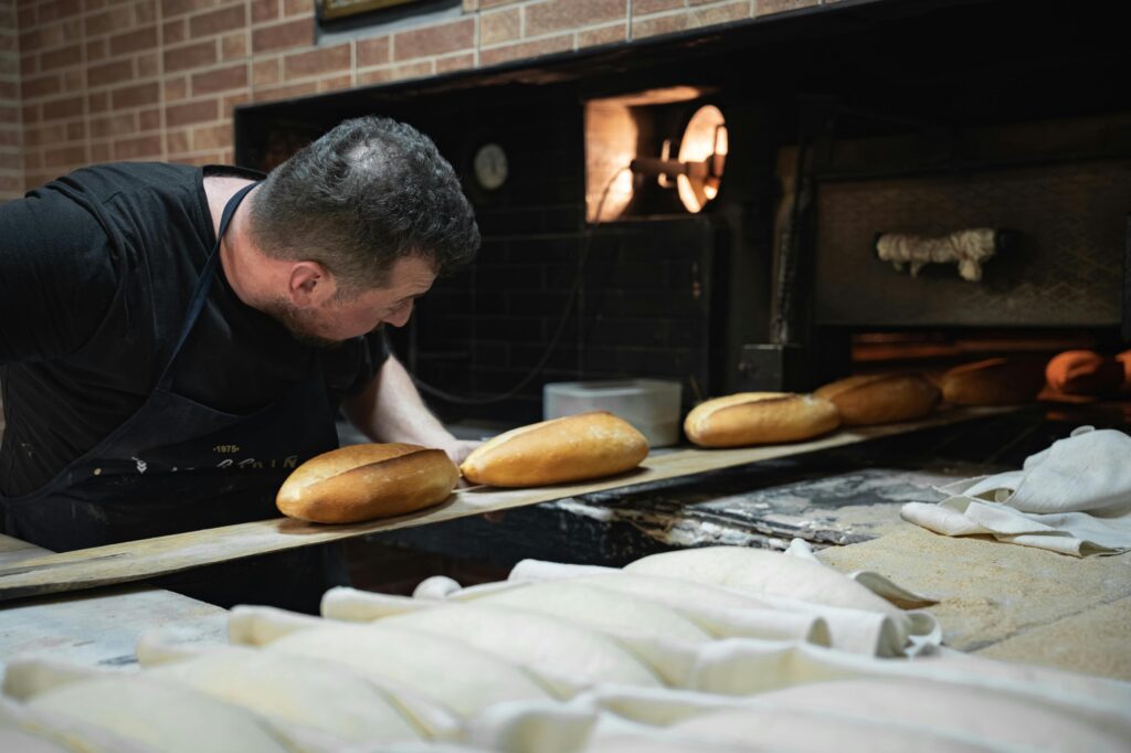 A baker leans over a long wooden peel, carefully sliding rows of crusty, golden-brown loaves out of a glowing stone oven. In the foreground, dozens of unbaked dough portions rest on floured cloths, waiting their turn for the heat.