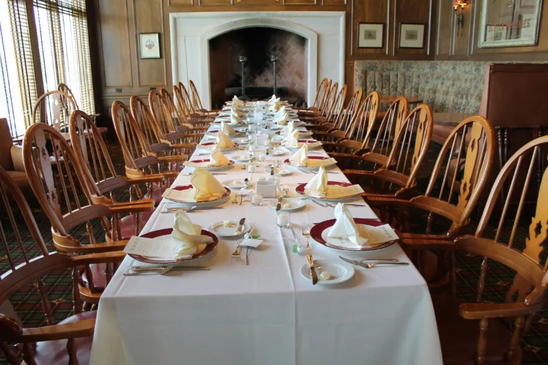 A long dining table is elegantly set with white linens, red-rimmed plates, and folded napkins in a rustic, wood-paneled room. High-backed wooden chairs line both sides of the table, which stretches toward a large stone fireplace at the far end.