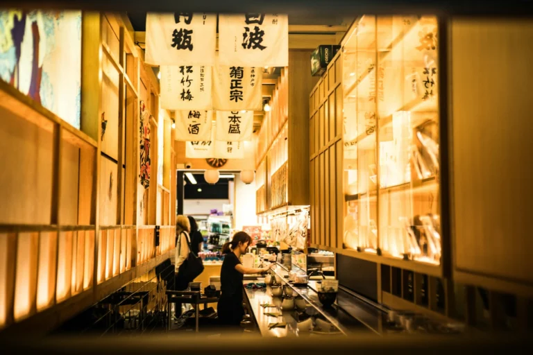 An eye-level shot framed by a dark foreground border captures the narrow, warmly lit corridor of a Japanese-style restaurant or market alley. White banners featuring bold black kanji hang from the ceiling above a woman working behind a long service counter lined with small dishes.