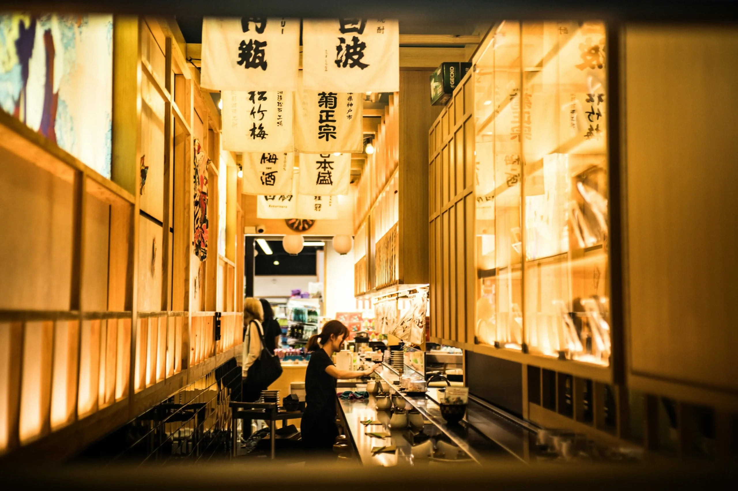 An eye-level shot framed by a dark foreground border captures the narrow, warmly lit corridor of a Japanese-style restaurant or market alley. White banners featuring bold black kanji hang from the ceiling above a woman working behind a long service counter lined with small dishes.