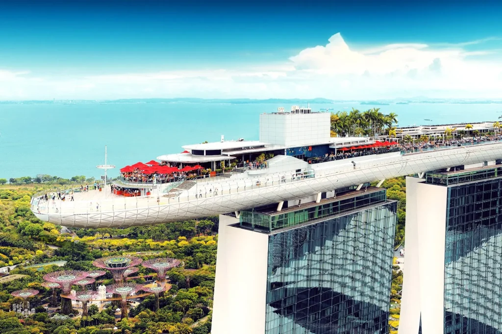 An aerial drone photograph showcases the iconic SkyPark cantilevered structure atop the Marina Bay Sands towers in Singapore, bustling with visitors and red umbrellas. In the lush green background below, the distinctive futuristic Supertree structures of Gardens by the Bay are visible against the ocean.