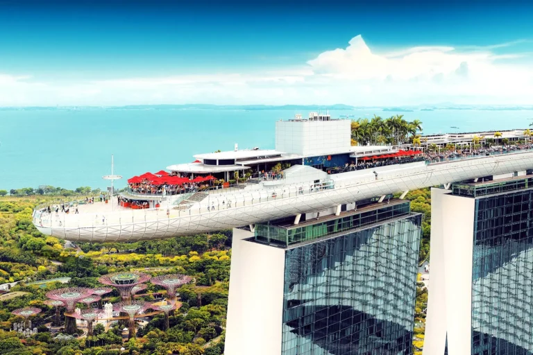 An aerial drone photograph showcases the iconic SkyPark cantilevered structure atop the Marina Bay Sands towers in Singapore, bustling with visitors and red umbrellas. In the lush green background below, the distinctive futuristic Supertree structures of Gardens by the Bay are visible against the ocean.