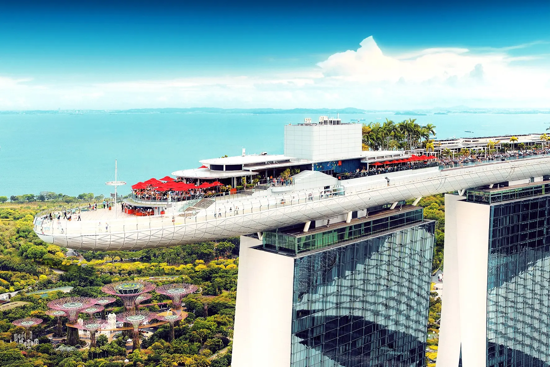 An aerial drone photograph showcases the iconic SkyPark cantilevered structure atop the Marina Bay Sands towers in Singapore, bustling with visitors and red umbrellas. In the lush green background below, the distinctive futuristic Supertree structures of Gardens by the Bay are visible against the ocean.