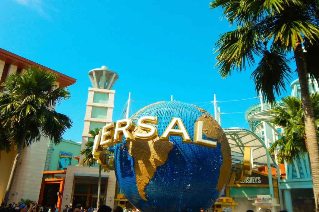 This image captures the iconic rotating Universal Studios globe located at the entrance of the theme park in Singapore. Surrounded by lush palm trees and modern architecture, the bright blue sphere stands out against a clear, sunny sky.
