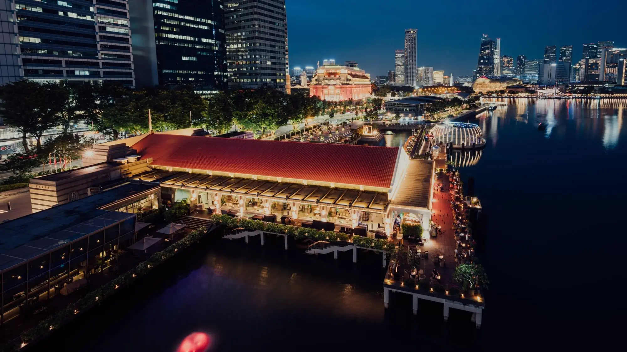 An aerial night shot captures a long, red-roofed building lit with warm lights along a dark waterfront, with people dining on an outdoor pier. In the background, a glowing city skyline stands against the dark night sky, reflecting beautifully on the surface of the water.