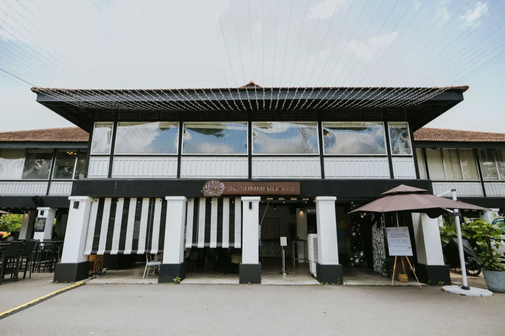 A two-story colonial-style building features a black and white facade with large windows and a brown tiled roof. Below a wooden sign reading "the SUMMERHOUSE," the open ground floor is decorated with striped awnings and string lights overhead.