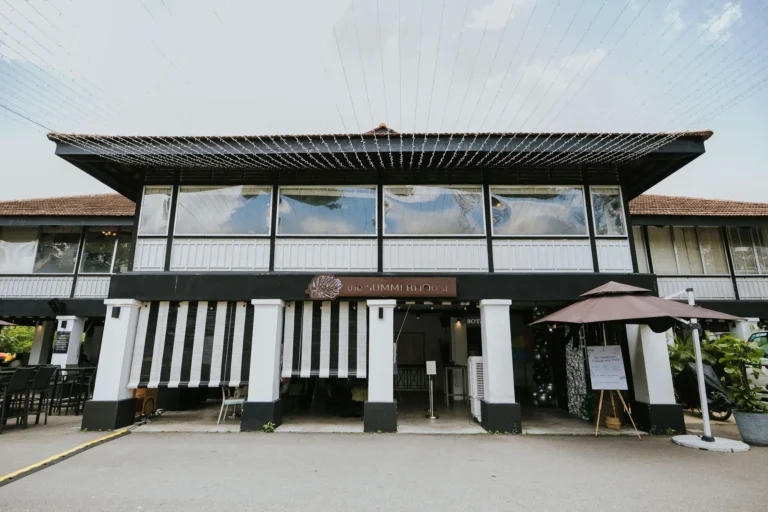 A two-story colonial-style building features a black and white facade with large windows and a brown tiled roof. Below a wooden sign reading "the SUMMERHOUSE," the open ground floor is decorated with striped awnings and string lights overhead.