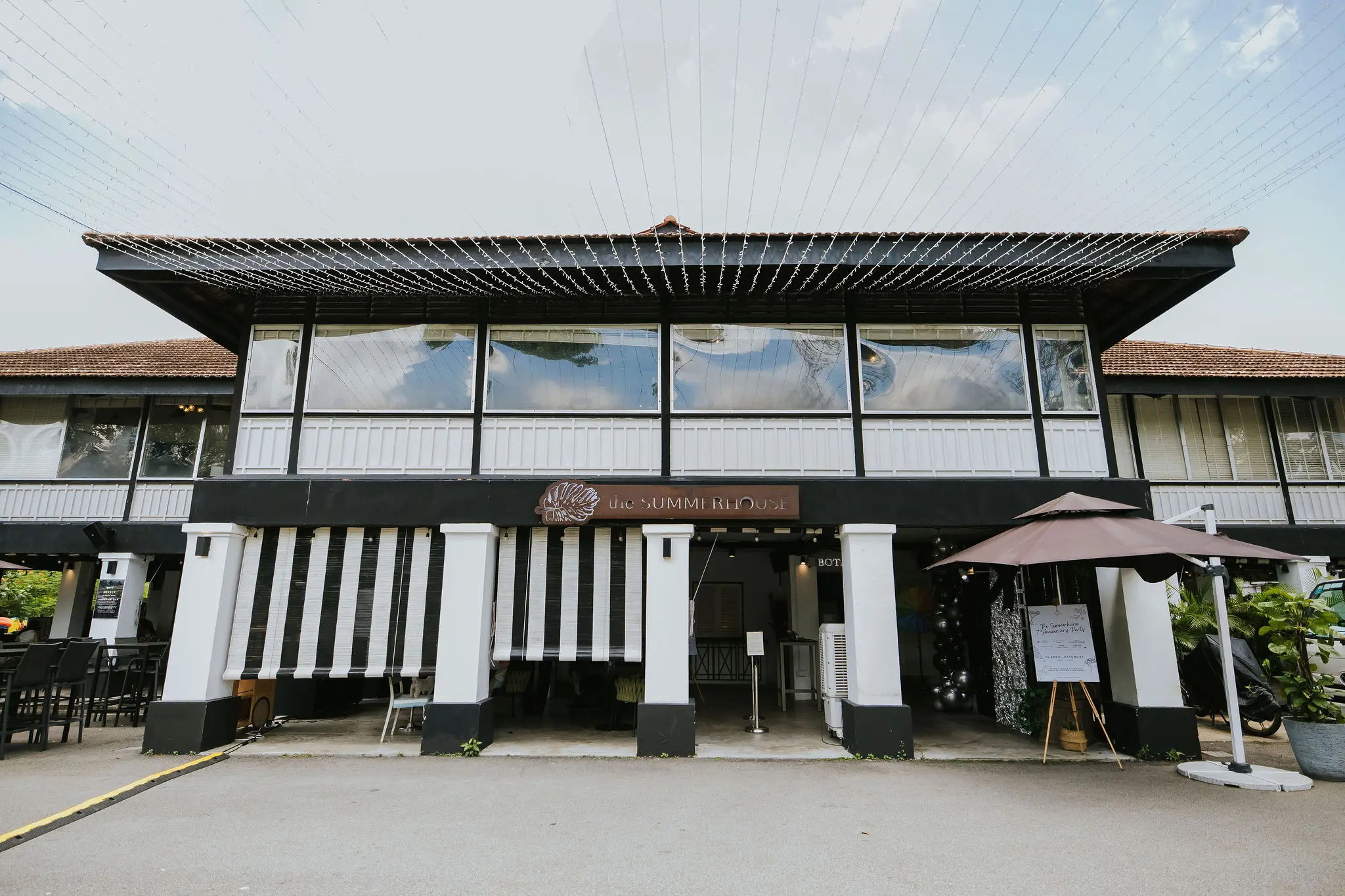 A two-story colonial-style building features a black and white facade with large windows and a brown tiled roof. Below a wooden sign reading "the SUMMERHOUSE," the open ground floor is decorated with striped awnings and string lights overhead.