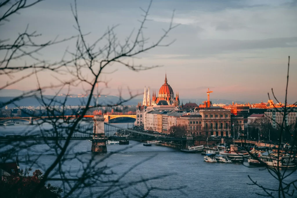 A scenic, high-angle view captures the Hungarian Parliament Building and the Danube River in Budapest during a warm sunset. Bare tree branches frame the foreground, looking past the Széchenyi Chain Bridge toward the city skyline under a soft, overcast sky.