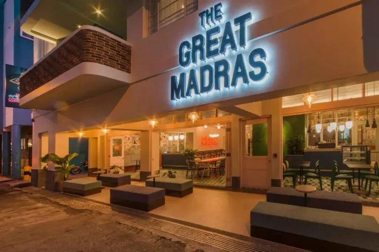 The facade of The Great Madras hotel glows with a vibrant blue neon sign above an inviting open-air seating area. The warm evening lighting showcases retro-inspired decor, including mid-century furniture and lush greenery tucked into the urban architecture.