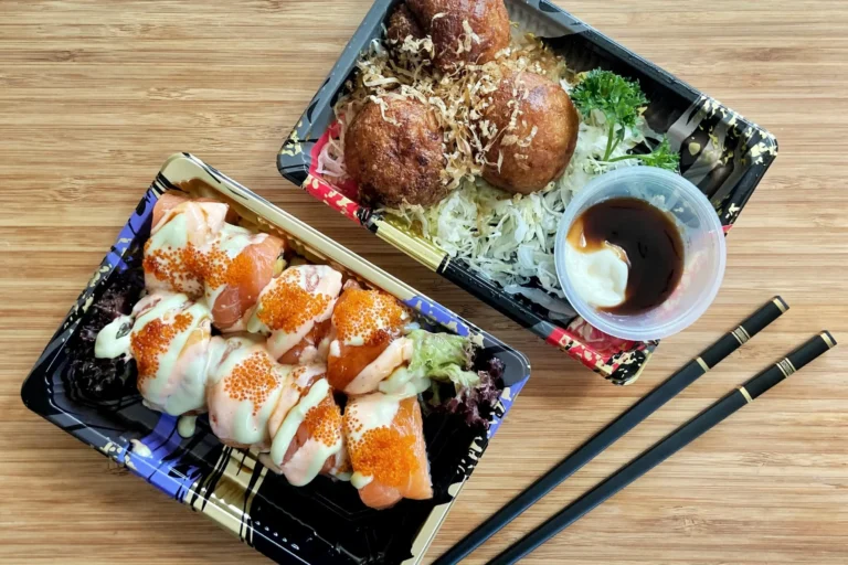 Two black plastic takeout containers filled with Japanese food sit on a light wooden table next to a pair of black chopsticks. The container on the left holds a salmon sushi roll drizzled with sauces and topped with orange fish roe, while the container on the right holds three golden-brown takoyaki balls served with shredded cabbage and a small cup of dipping sauce.