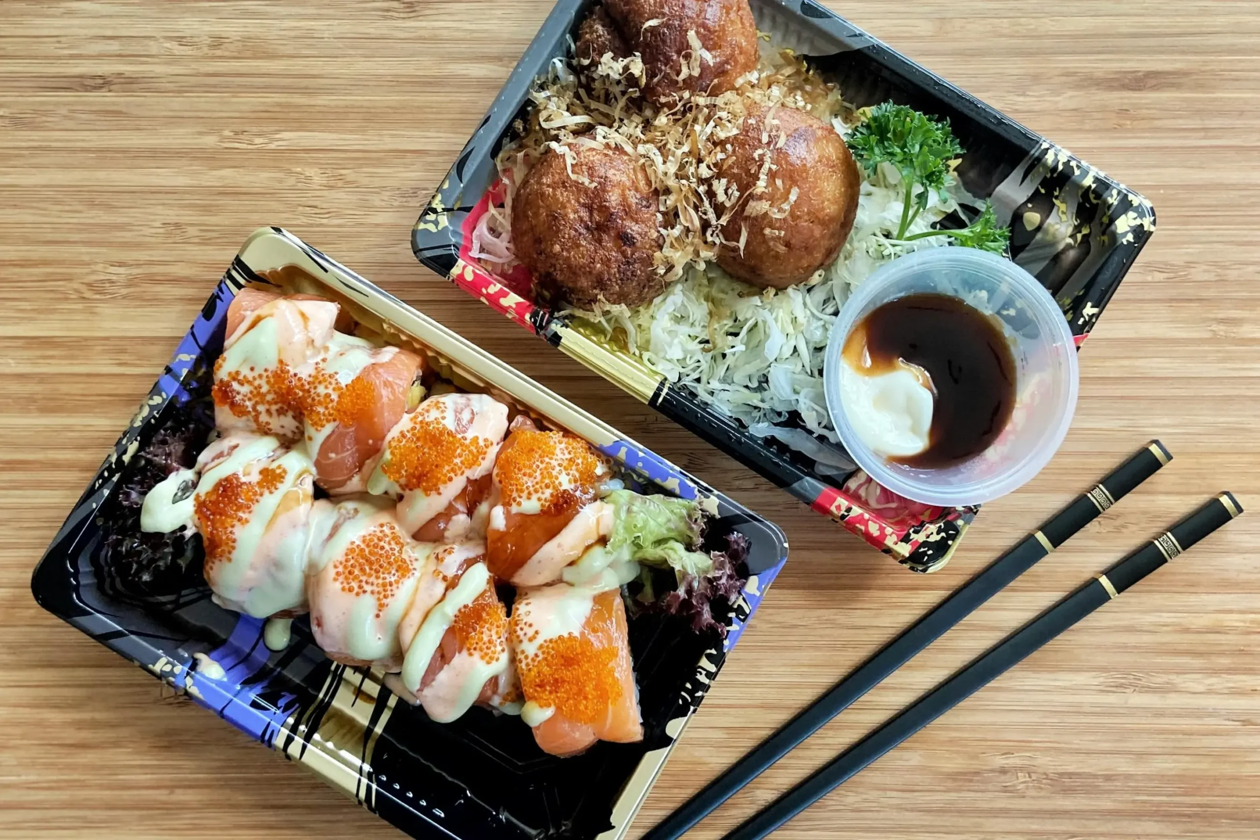 Two black plastic takeout containers filled with Japanese food sit on a light wooden table next to a pair of black chopsticks. The container on the left holds a salmon sushi roll drizzled with sauces and topped with orange fish roe, while the container on the right holds three golden-brown takoyaki balls served with shredded cabbage and a small cup of dipping sauce.