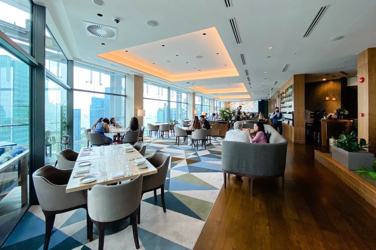 The image shows a modern, upscale restaurant with large floor-to-ceiling windows offering a panoramic view of a city skyline. The interior features a distinctive geometric-patterned carpet, warm recessed lighting, and several patrons seated at tables.
