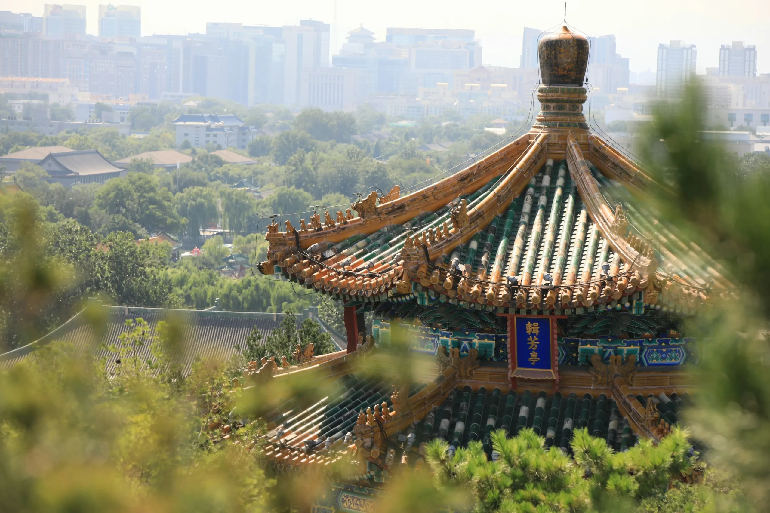 This image features an intricate, multi-tiered traditional Chinese pavilion with a vibrant green-tiled roof and ornate gold carvings. In the background, a hazy modern city skyline creates a striking contrast with the ancient architecture and lush surrounding greenery.