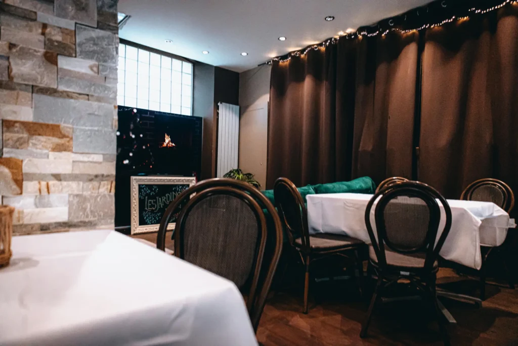 This image shows a dimly lit indoor dining area featuring tables set with white tablecloths and dark wooden chairs with rounded backs. Large brown curtains and a stone-textured wall create a cozy atmosphere, while a small fireplace glow is visible in the background.