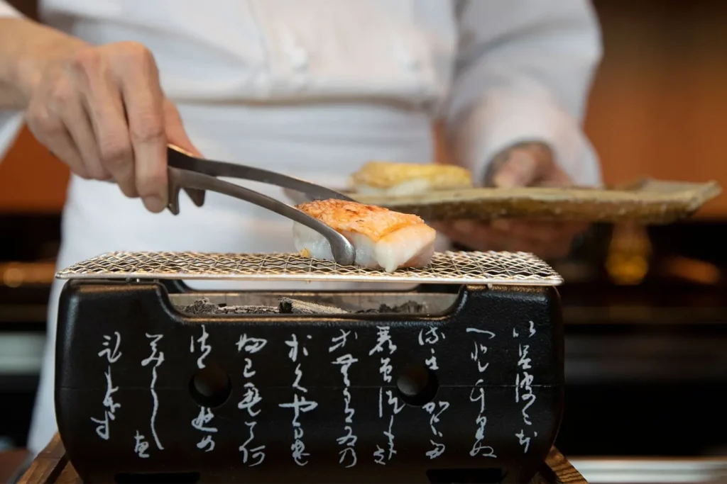 A chef uses metal tongs to carefully place a piece of fish with crispy, seared skin onto a small tabletop charcoal grill. The black ceramic grill features elegant white Japanese calligraphy inscribed across its front surface.