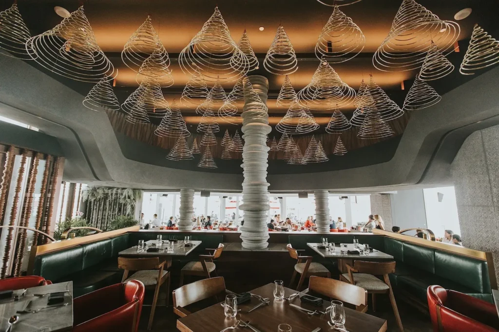 An upscale restaurant dining room features dark green leather booths and wooden tables arranged beneath a striking ceiling installation of numerous wire, spiral-coned sculptures. A central, segmented white stone pillar anchors the space, while large windows in the background reveal an outdoor patio with patrons.