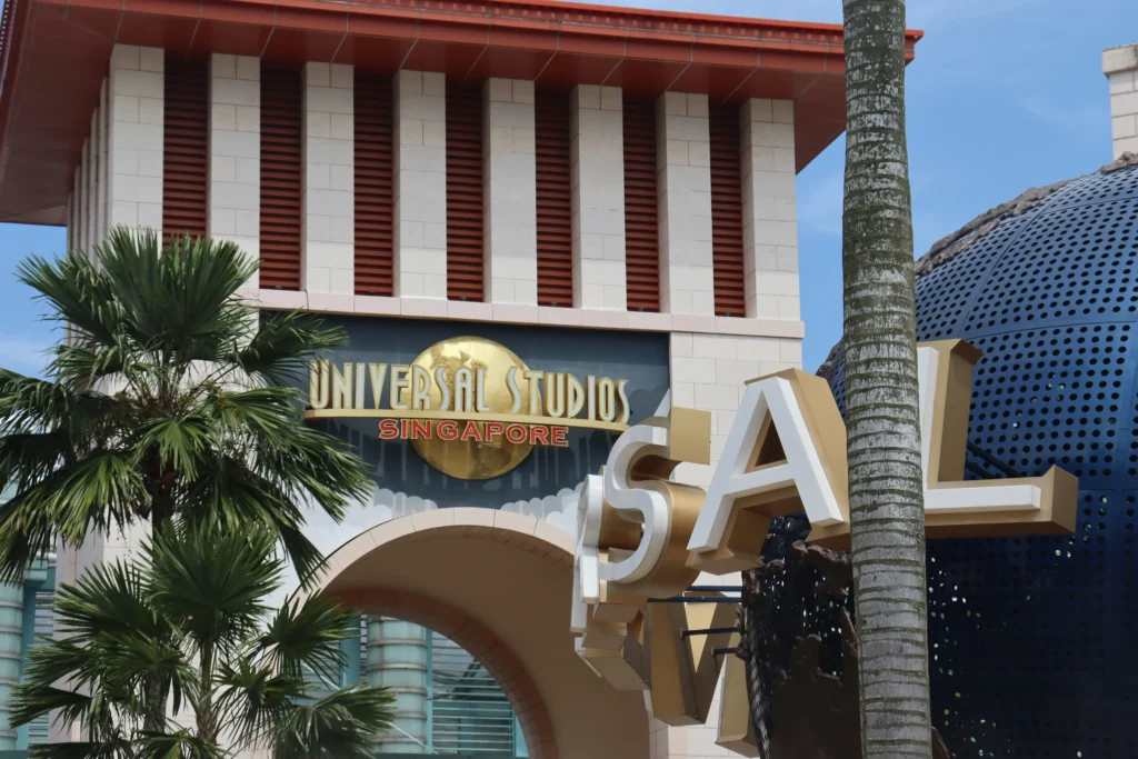 The iconic Universal Studios Singapore logo is displayed prominently on a tan, architectural archway framed by a tropical palm tree. In the foreground, the large, metallic letters of the famous rotating globe catch the sunlight against a clear blue sky.