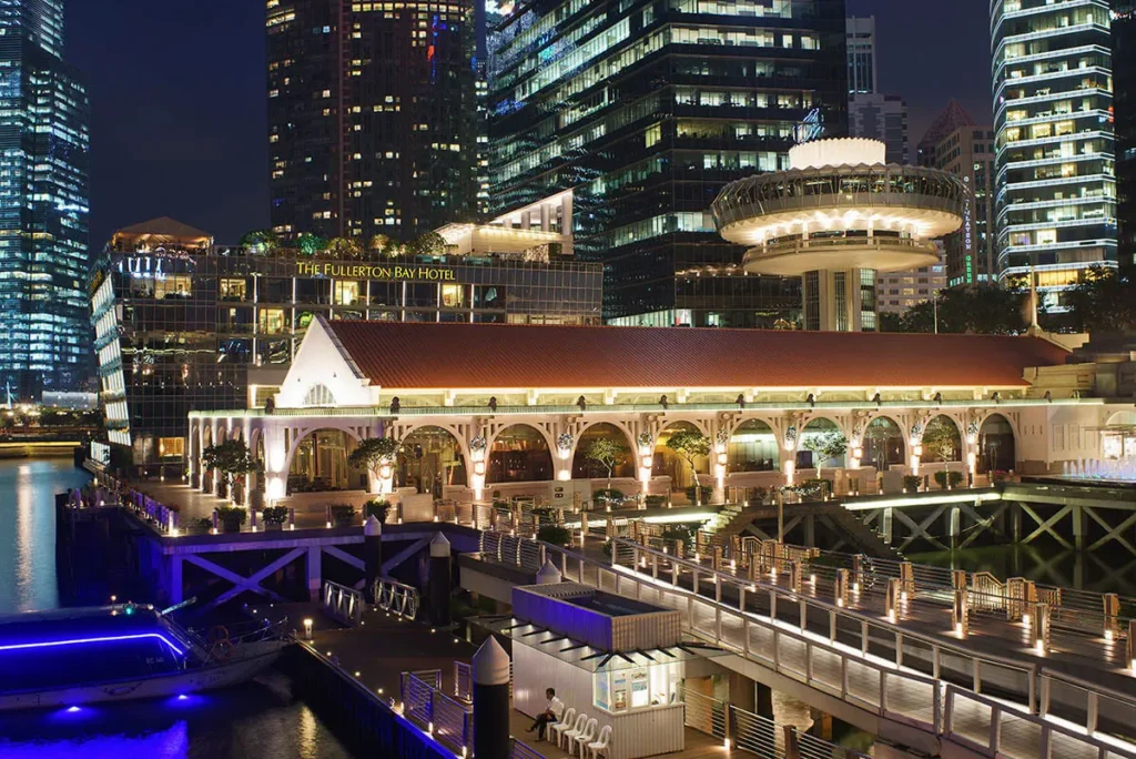 A night shot captures the illuminated, historic Clifford Pier building with its red roof and arched facade, situated on the waterfront in front of modern skyscrapers and The Fullerton Bay Hotel. In the foreground, a lit pedestrian walkway stretches across the water, where a small boat with blue lights is docked.