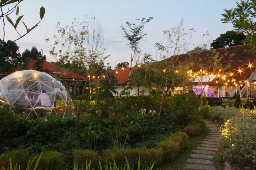 Two people sit at a table inside a glowing geodesic garden dome at dusk, surrounded by lush greenery and a stone pathway. In the background, warm string lights illuminate a traditional building with a terracotta roof under a soft evening sky.