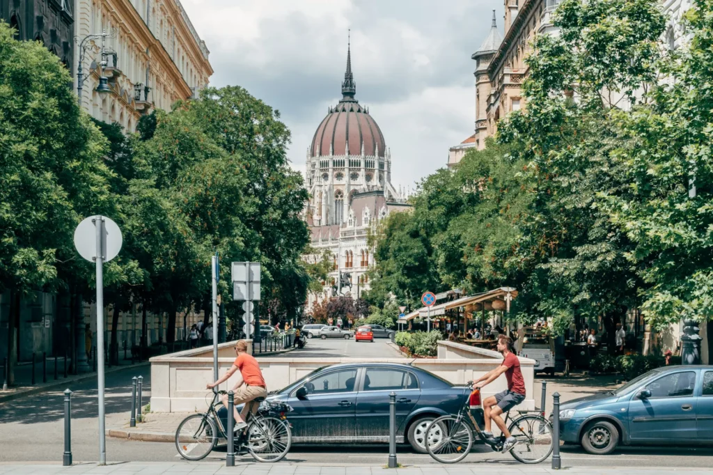A street view captures two cyclists riding past a dark sedan on a tree-lined city street. In the background, the grand, domed structure of the Hungarian Parliament Building rises prominently under a cloudy sky.