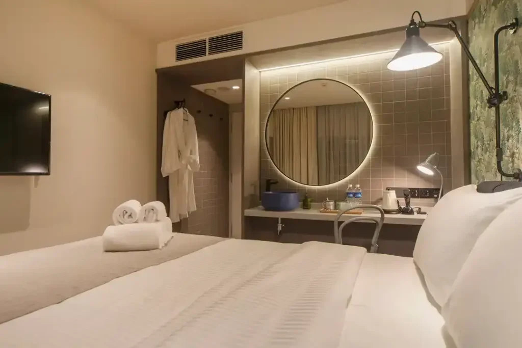 This modern hotel room features a crisp white bed in the foreground and a vanity area with a large circular backlit mirror. A white bathrobe hangs in an open closet nook, while industrial-style lighting and neutral tones create a clean, contemporary atmosphere.