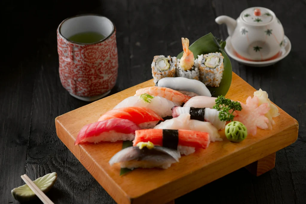 A traditional Japanese sushi platter featuring an assortment of nigiri and maki rolls is beautifully arranged on a wooden *geta* board. The setting is completed with a cup of green tea, a small teapot, and chopsticks on a dark wooden table.