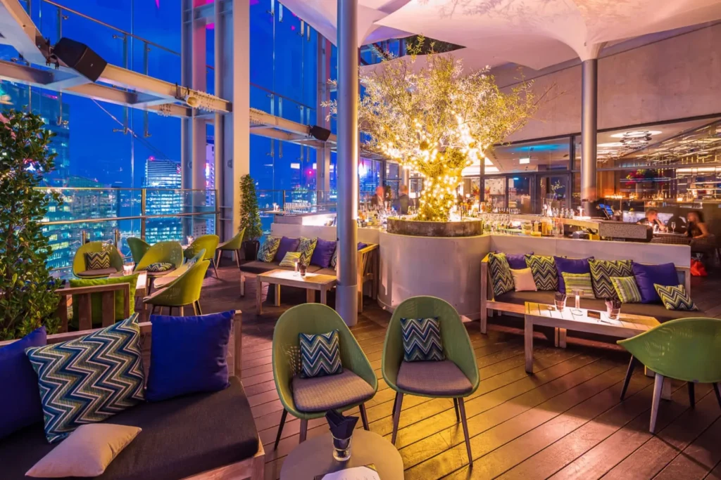 The image depicts a chic rooftop lounge featuring vibrant green and blue seating arranged around an illuminated tree. Large glass walls offer an expansive view of a glowing city skyline under a deep twilight sky.