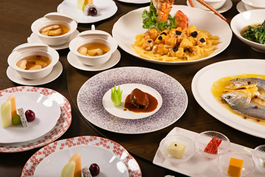 An elegant Chinese banquet is meticulously arranged on a dark wooden tabletop, showcasing a variety of high-end Cantonese dishes. The spread features steamed whole fish in a yellow broth, braised abalone with bok choy, and lobster pasta accented with black truffles.