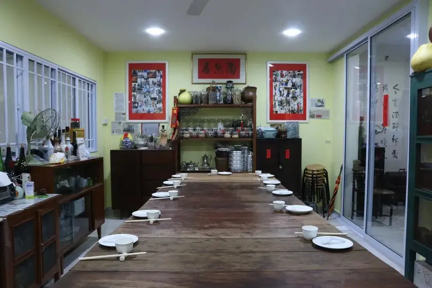 A long, rustic wooden table is set for a group meal with white bowls and chopsticks in a room with light yellow walls. The background features shelves filled with jars, traditional Chinese decor, and various pieces of framed artwork.