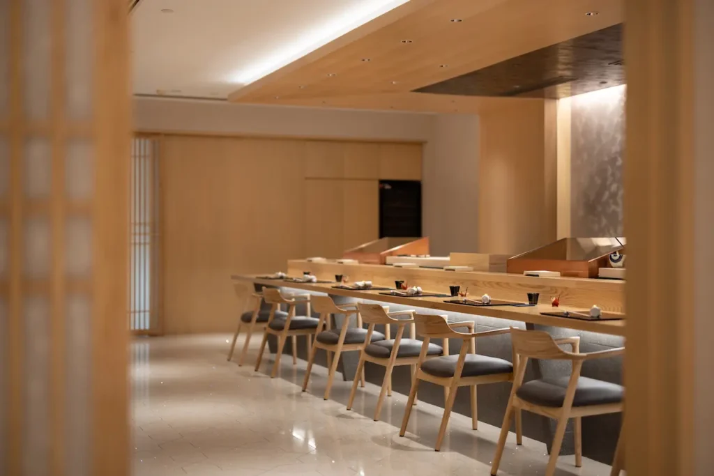An elegant, minimalist Japanese counter-service restaurant features a long wooden bar lined with sleek wooden armchairs with grey upholstered seats. The warm-toned interior is highlighted by soft recessed lighting, light wood paneling, and meticulous place settings awaiting guests.