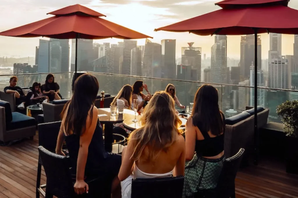 A group of women sit at a table on an outdoor rooftop terrace, enjoying drinks under large red umbrellas. In the background, a sprawling cityscape of modern skyscrapers is illuminated by the warm glow of a setting sun.