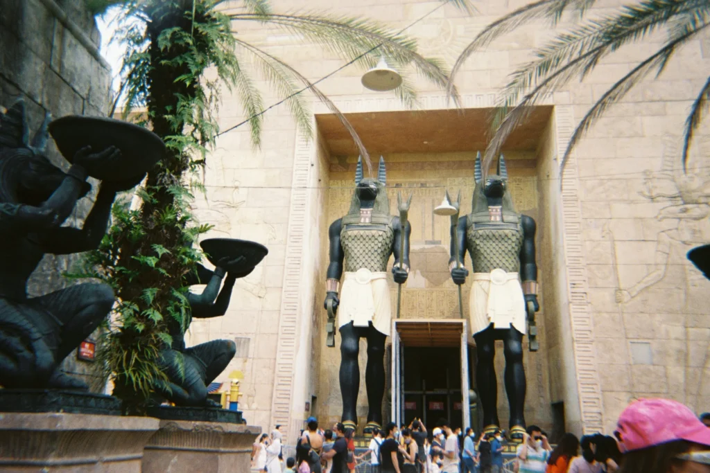 Two towering statues of Anubis flank a large, temple-like entrance adorned with Egyptian motifs and hieroglyphics. A crowd of people gathers at the base of the structure, which is framed by lush palm trees and stone sculptures.