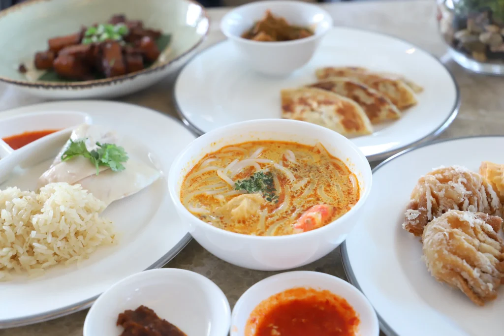A bowl of creamy, orange laksa soup with noodles sits at the center of a table filled with various Southeast Asian dishes. Surrounding the soup are plates of Hainanese chicken rice, roti canai with curry dipping sauce, braised pork, fried dumplings, and small bowls of chili sauce.