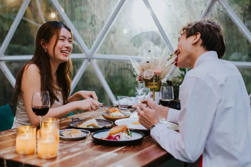 A smiling couple enjoys a romantic dinner inside a glass garden dome, surrounded by warm candlelight and several gourmet dishes. They share a joyful conversation over glasses of red wine while seated at a rustic wooden table decorated with a floral centerpiece.