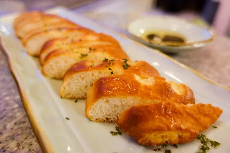 Slices of toasted bread with golden-brown crusts are arranged in a row on a long, rectangular white plate. Small green herbs are sprinkled over the bread, and a small dish of oil and balsamic vinegar sits in the blurred background.