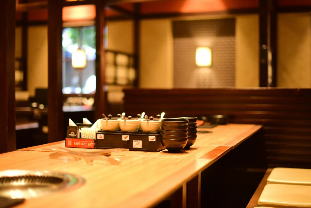 A wooden dining table inside a restaurant is neatly set with small condiment jars, stacks of black bowls, and a built-in tabletop grill. The background shows a warmly lit, cozy interior with wooden partitions and soft ambient lighting.