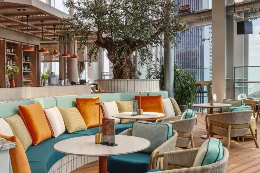 The image displays an upscale rooftop lounge with colorful seating and a large decorative tree as a focal point. Modern skyscrapers are visible through the glass perimeter, while warm lighting and plush cushions create a welcoming atmosphere for dining or drinks.