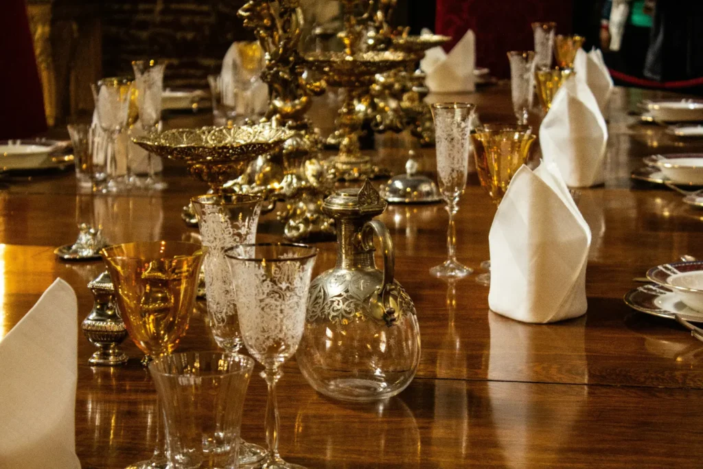 An elegant, long wooden dining table is meticulously set with ornate glassware, golden centerpieces, and decorative silver decanters. Crisp white napkins are folded into tall peaks beside intricate place settings, creating a grand and formal atmosphere.