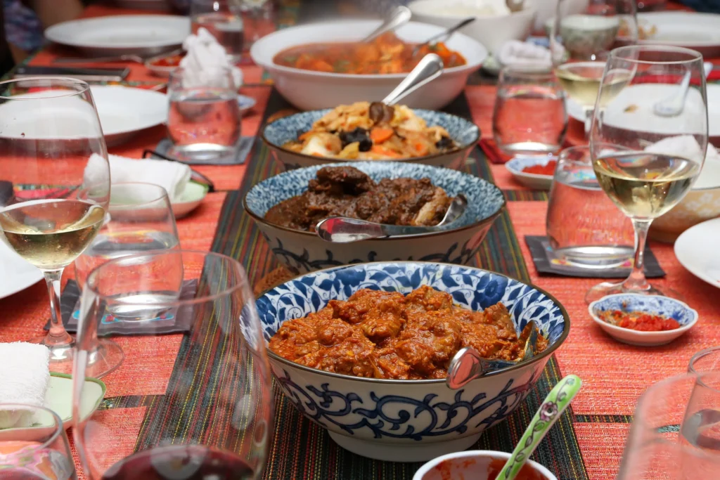 A series of ornate blue-and-white bowls filled with colorful Peranakan-style dishes is arranged down the center of a long dining table. The setting is formal and communal, featuring wine glasses, water tumblers, and patterned runners laid over a vibrant orange tablecloth.