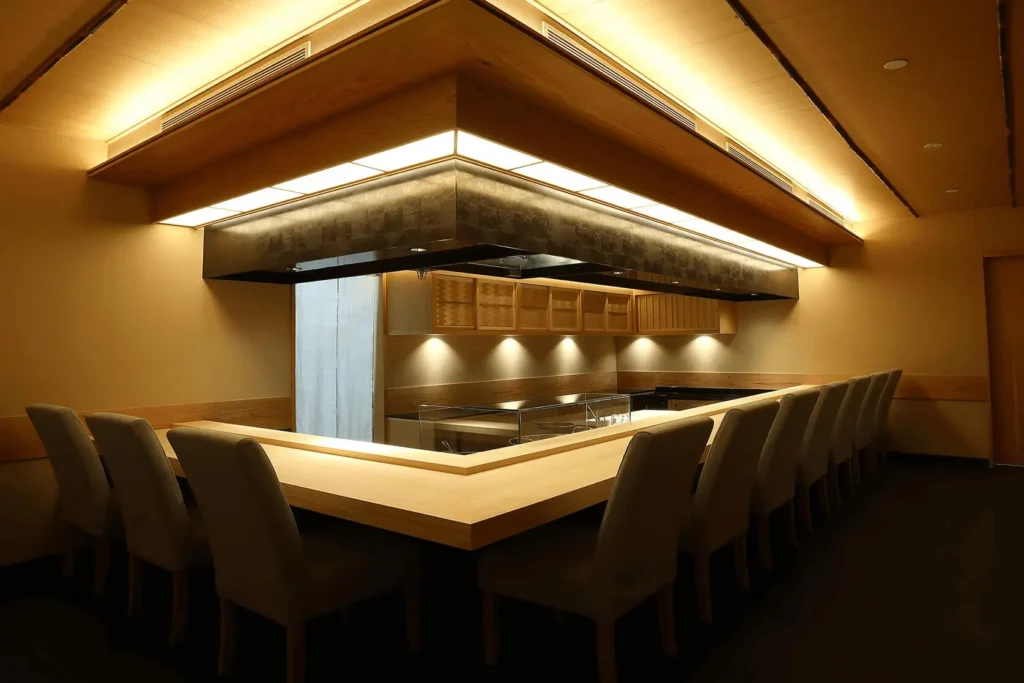 A minimalist, high-end Japanese sushi counter features a light-colored wooden L-shaped bar surrounded by elegant upholstered chairs. Warm, indirect lighting glows from the coffered wooden ceiling and accentuates the sleek dark metal exhaust hood above the chef's preparation area.