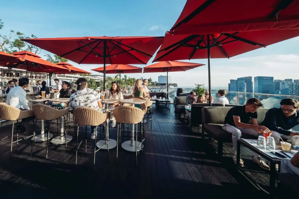 A lively rooftop bar features people seated at tables and lounges under large, bright red umbrellas on a sunny day. The dark wooden deck overlooks a modern city skyline with skyscrapers visible in the background.