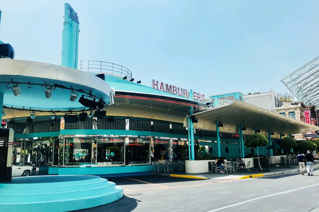 This image features a retro-themed "Mel's Drive-In" diner with a distinctive turquoise and white Googie-style architecture. The scene captures the classic 1950s aesthetic, complete with a circular driveway, neon signage, and outdoor seating under a wide, sweeping canopy.