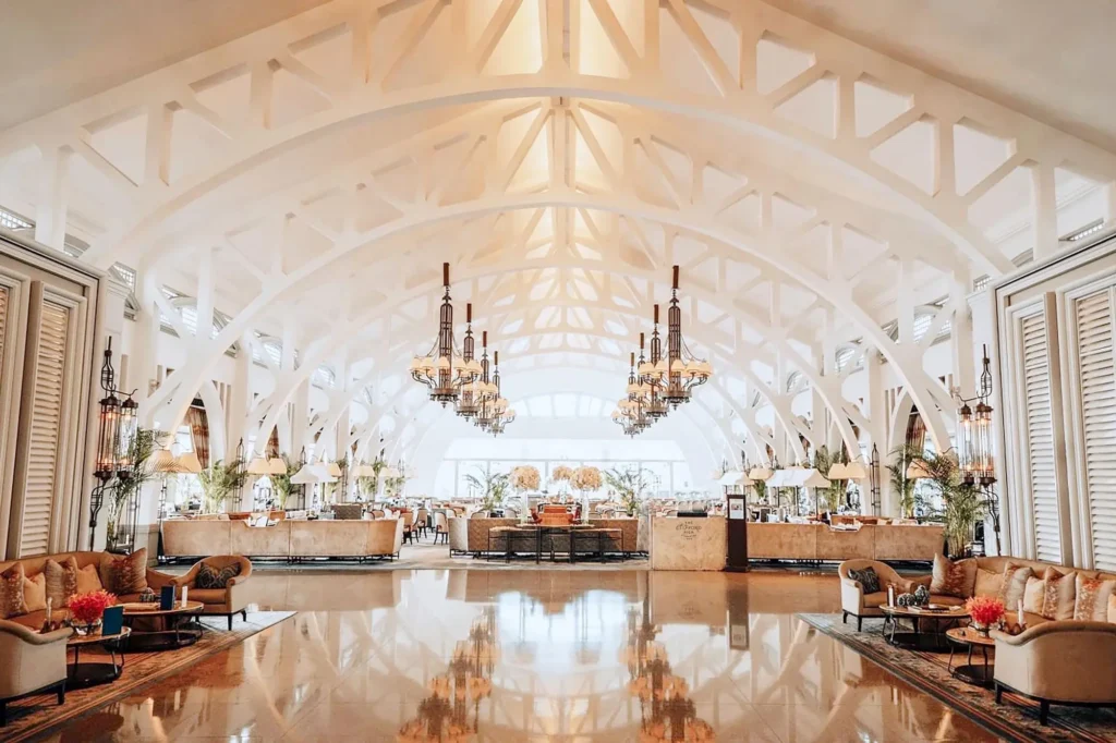 A spacious, elegant hall features a high, vaulted ceiling with white arched trusses and polished floors that reflect the bright, warm lighting. Intricate multi-tiered chandeliers hang above symmetric arrangements of plush sofas, armchairs, and tables, creating a luxurious and inviting lounge atmosphere.
