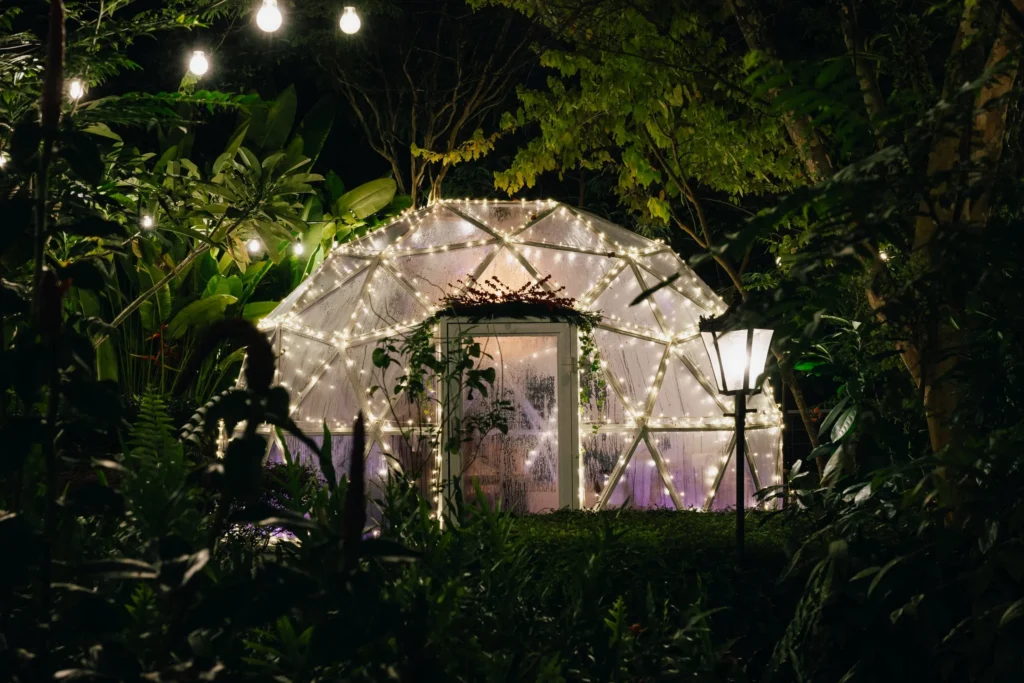 A glowing, transparent geodesic dome sits nestled within a lush, dark garden at night, decorated with strings of warm white lights along its frame. The surrounding tropical foliage is softly illuminated by hanging bistro lights and a vintage-style lamp post in the foreground.